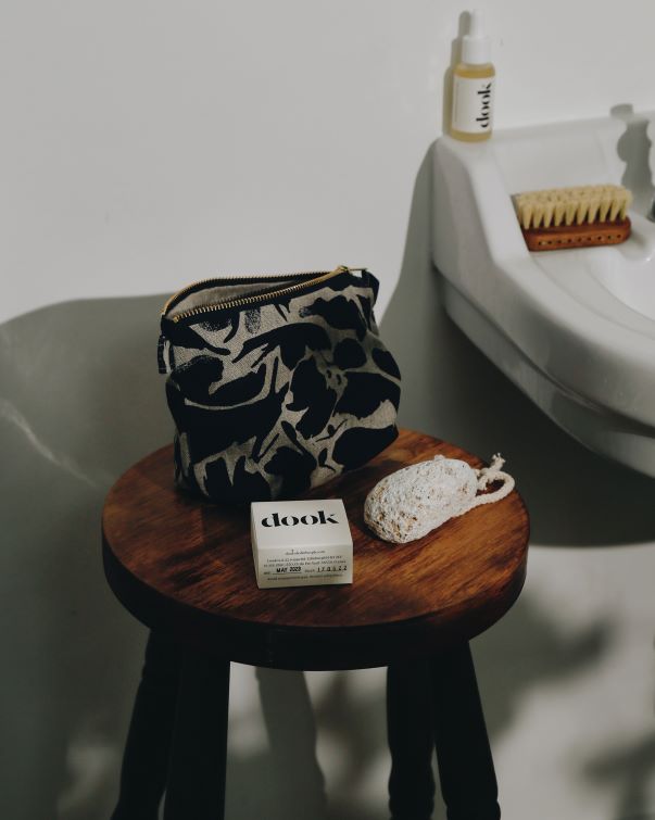 Linen cosmetic bag with irregular dark navy shapes standing on the wooden stool next to a shampoo bar and a pumice stone next to the sink in the bathroom.