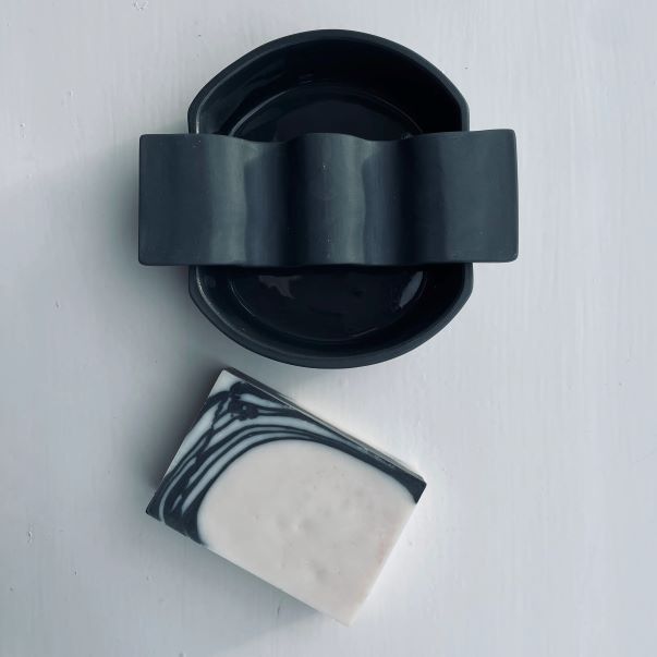 Ceramic soap dish with a wavy part on the top with a rosemary black patterned handmade soap on the white background, shot from the top.