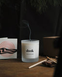 Dook candle in a cosy room freshly blown and releasing smoke. Piece of candle packaging, matches used as decor and some pine needles hanging above. Entire composition arranged in a dark cosy room. 