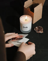 Hands folding candle packaging into a card next to a lid candle and empty brown box.