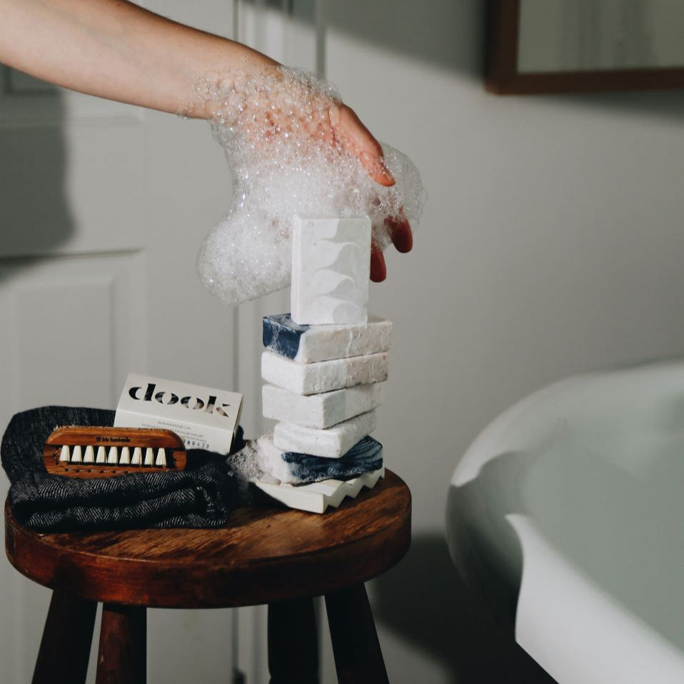 A foam covered hand supporting the stack of soaps placed on the soap and a wooden stool standing next to a bath tub.