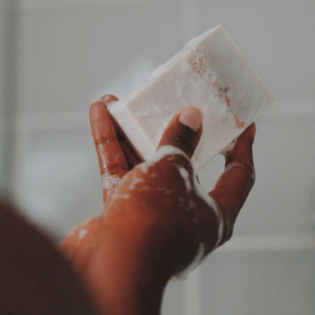 A hand holding a bar of wet and lathered soap on the white blurred bathroom tiles background.