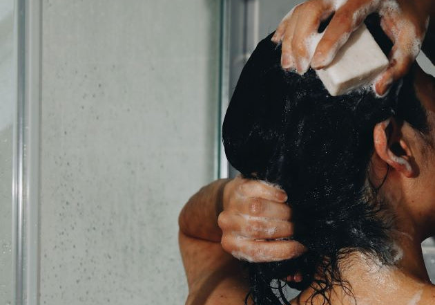 A black haired woman turned back lathering up the shampoo bar under the shower.