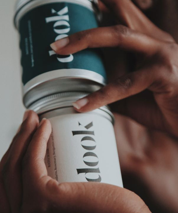 Two hands holding together two different tins of dook bath salts.