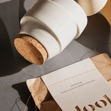 A shoot from the top of white ceramic jar with cork lid laying next to mandarin bag of bath salts. 