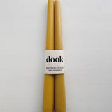 A pair of yellow, beeswax dining candles wrapped with natural creamy paper with black dook logo.