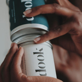 Two differently labelled tins with bath salts. One on tops of the other, being hold by two hands on the blurred body background.