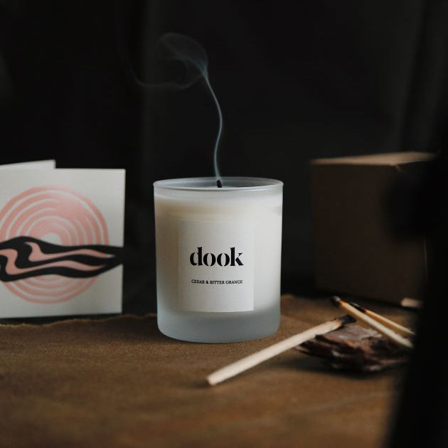 Dook candle in a cosy room freshly blown and releasing smoke. Piece of candle packaging, matches used as decor and some pine needles hanging above. Entire composition arranged in a dark cosy room. 
