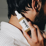 Profile of black haired man with beard holding a bottle of conditioning oil and presenting it on his neck.