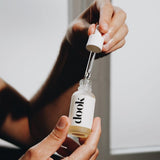 Hands holding small glass bottle of conditioning oil in one hand and a pipette in the other hand, right above the bottle. Black dook logo on the white label stick to the bottle.