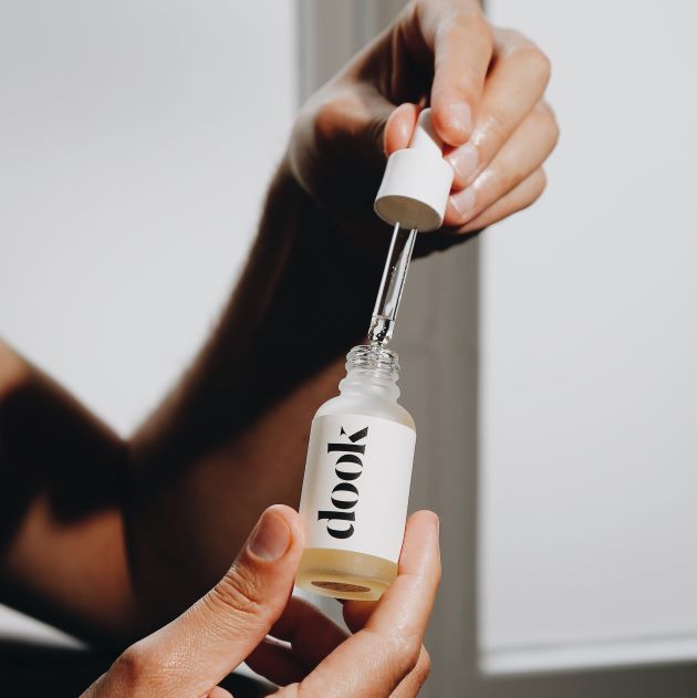 Hands holding small glass bottle of conditioning oil in one hand and a pipette in the other hand, right above the bottle. Black dook logo on the white label stick to the bottle.