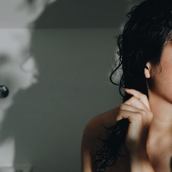 A woman in the bathroom holding her damp long black hair applying conditioning oil.