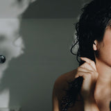 A woman in the bathroom holding her damp long black hair applying conditioning oil.