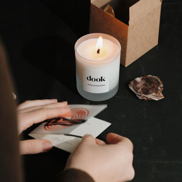 Hands folding candle packaging into a card next to a lid candle and empty brown box.