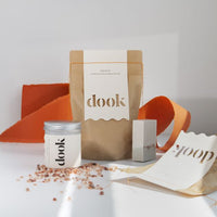 A composition of mandarin scented bath salts in two sized packaging. Two brown bags labeled with logo on white paper and aluminium tin with black logo on white paper. A handful of himalayan rock salt spread on the surface and orange torn paper in the background and matching scent soap standing next to a bag. All on white background.. 