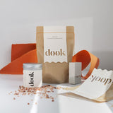 A set of dook products - bath soak tin and bag standing next to a bar of soap. An orange torn piece of paper create a decorative background/ Raw pink salt crystals sprinkled on the surface.