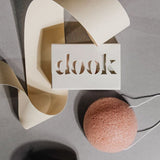 An artistic arrangement of a dook soap in a packaging lying flat on grey surface next to a round pink konjac sponge. 
