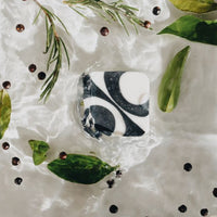 A black and white, square-shaped handmade bar of soap placed flat in a water with floating leaves and juniper dried fruits on the white background with shades being reflected from the floating leaves..