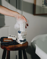 A foam covered hand supporting the stack of soaps placed on the soap and a wooden stool standing next to a bath tub. 