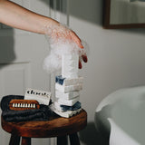 A foam covered hand supporting the stack of soaps placed on the soap and a wooden stool standing next to a bath tub. 