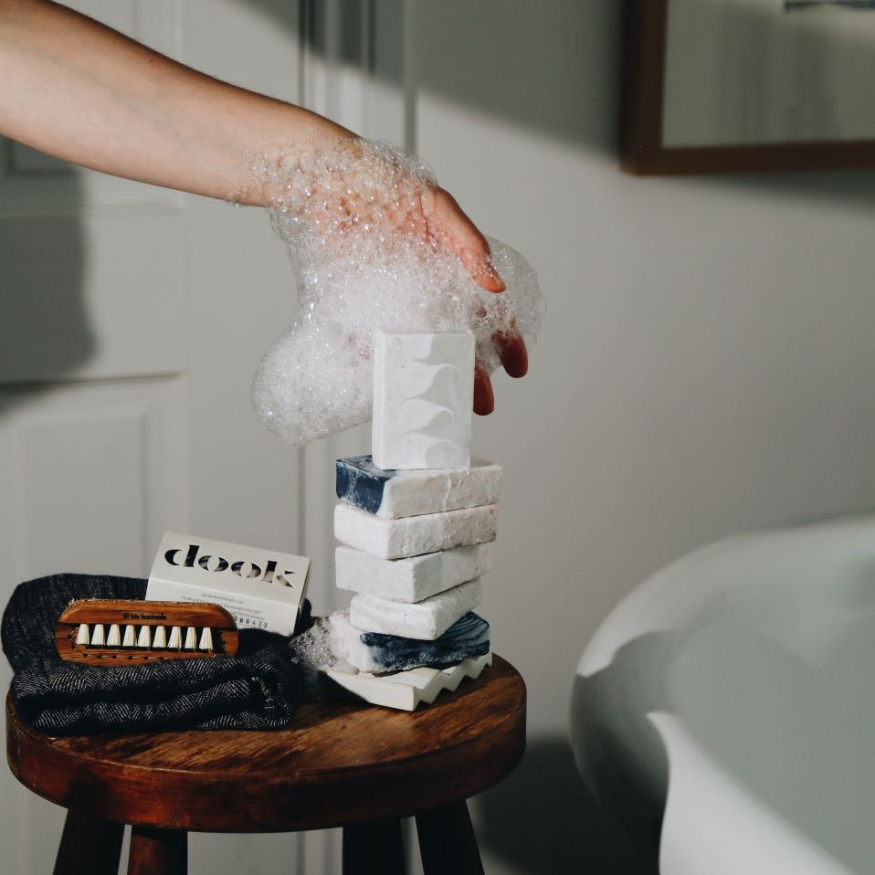 A foam covered hand supporting the stack of soaps placed on the soap and a wooden stool standing next to a bath tub. 