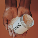 Two hands open holding a handful of epsom bath salts and an open aluminium tin with salts inside and outer white label with black dook logo all on copper orange bacground.