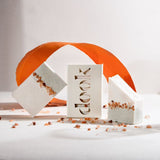 Three bars of handmade salt soap arranged in an arty chaotic composition. Bare soap and soap in a box with the dook logo surrounded by a decoration of pink, sprinkled around salt rocks, and an orange torn piece of paper shaped in a round way, all on the white background.