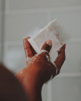 A hand holding a bar of wet and lathered soap on the white blurred bathroom tiles background.