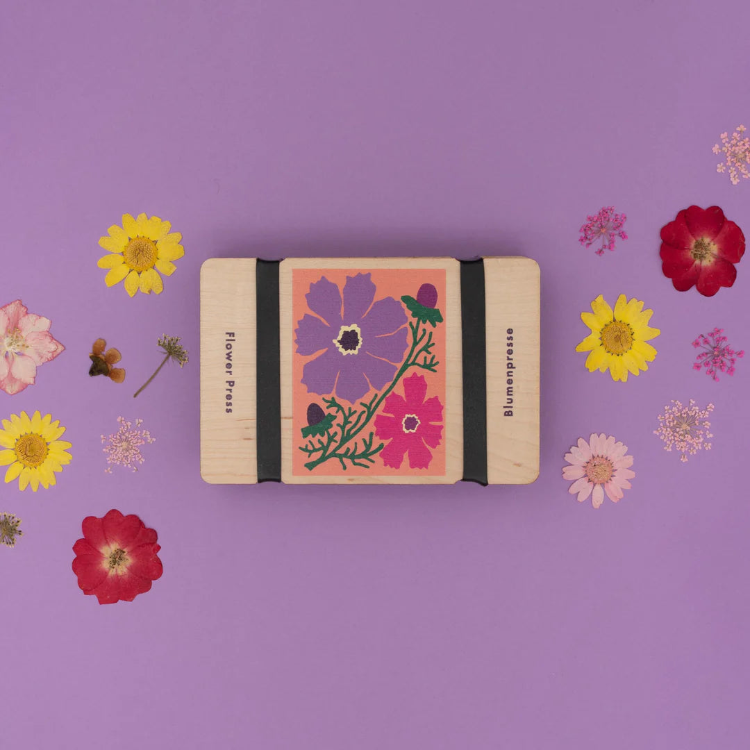 A pocket-sized flower press with a floral design, surrounded by various dried flowers and leaves on a purple background.