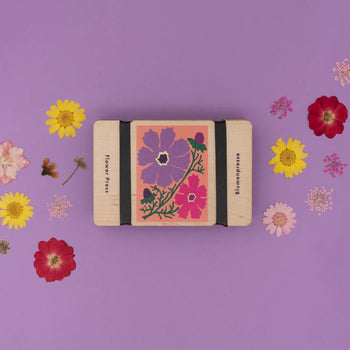 A pocket-sized flower press with a floral design, surrounded by various dried flowers and leaves on a purple background.