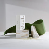 Stack of three bar soaps, two without packaging laying horizontally with one in a box placed vertically on the top. Green piece of torn paper behind acting as a leaf and a broken piece of another soap next to the stack.