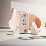 An arty composition of one boxed and two unboxed soaps stacked horizontally and vertically with one broken soap on the background including torn pink paper and shades on the white background.