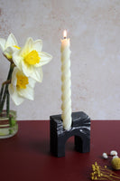 A twisted tall candle placed in a stone black and white candle holder standing on the red table next to daffodils.