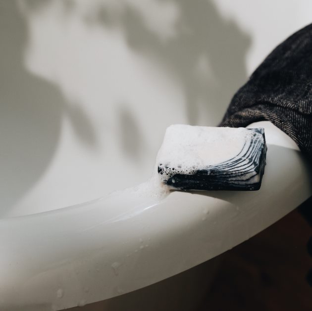 A handmade bar of soap and piece of a towel placed on the rim of a bath tub. Plant shades in the background.