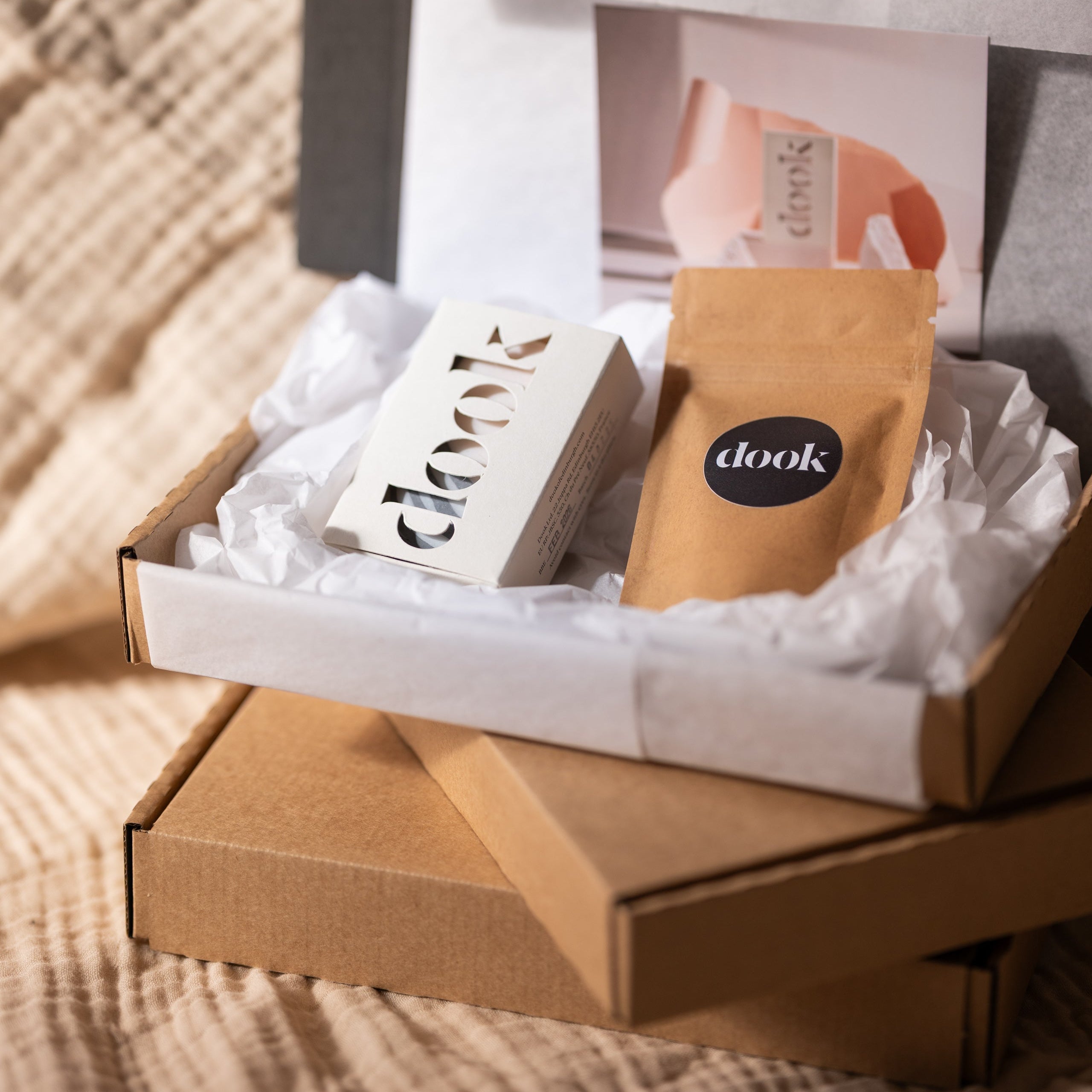 An open gift box containing a scented handmade soap bar, a small sample packet, and a handwritten note, all presented on a soft surface.