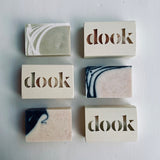 Three boxed and three unboxed dook soaps lying flat on the white surface in two columns of three soaps, exposing variety of soap patterns and logo on the box.