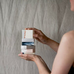 A women holding a stack of six different dook bar soaps on the background of a linen curtain.