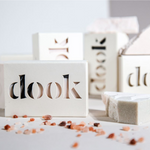 Dook soaps in packaging of a white box with a logo arranged chaotically with blurred bacground of soaps standing vertically behind. A broken in half light green soap on the right and some other broken soaps blurred in the background. Himalayan pink rock salt it the front sprinkled irregularly. 