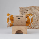 A yellow print patterned linen eye pillow on a wooden bridge block with a tree bark in the background.