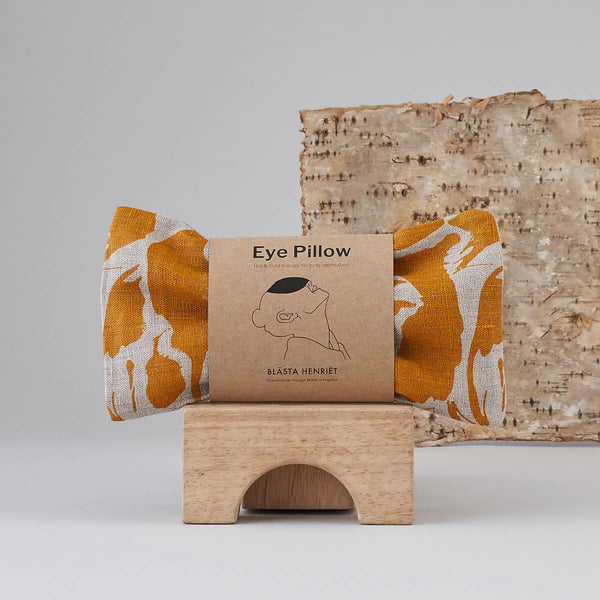 A yellow print patterned linen eye pillow on a wooden bridge block with a tree bark in the background.