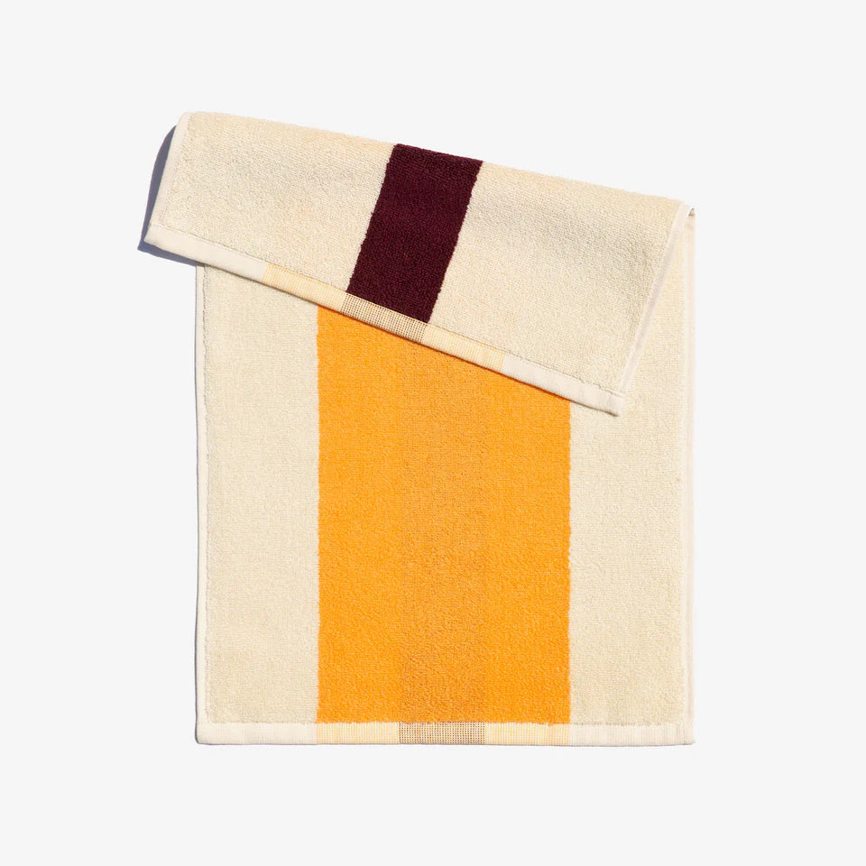 A folded face towel featuring a geometric pattern with amber and bordeaux stripes on a cream background, displayed neatly with a loop on the top corner.