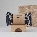 An navy print patterned linen eye pillow placed on a wooden bridge block with a tree bark in the background. 