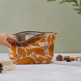 A hand exposing the inside of the yellow print patterned wash bag. Composition on the green bacground with a cosmetic brushes, soaps and a book standing next to a bag.