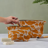 A person's hand putting a make up brush into a smaller, yellow print patterned linen wash bag. Bigger size behind on the green background. Unboxes soaps and another make up brush in the photo arrangement.