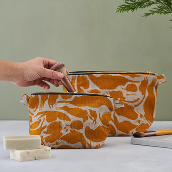 A person's hand putting a make up brush into a smaller, yellow print patterned linen wash bag. Bigger size behind on the green background. Unboxes soaps and another make up brush in the photo arrangement.